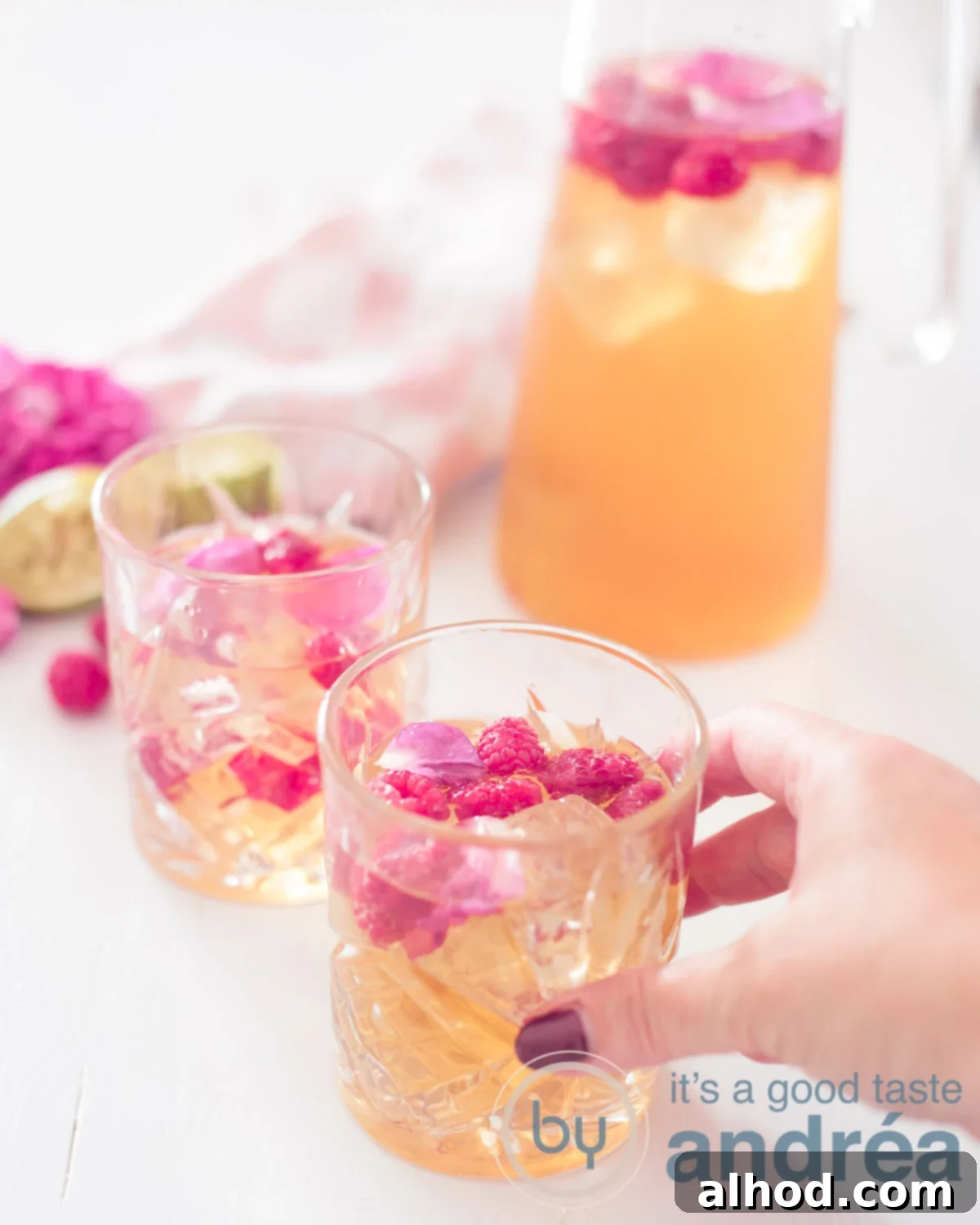 Aromatic Rose & Zesty Lime Iced Tea 5 Two glasses filled with iced tea with lime, rose and raspberries on a white background. A decanter in the background. A hand takes the front glass, ready to enjoy.