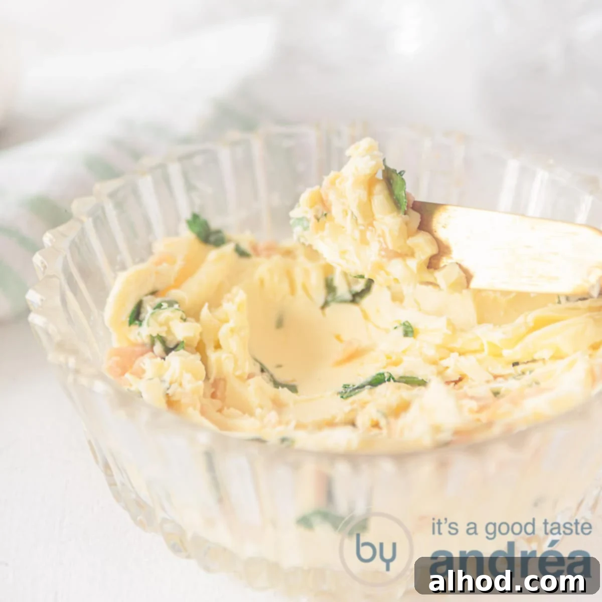 A square photo with a glass bowl filled with easy garlic butter. A gold knife takes some off. A white background.