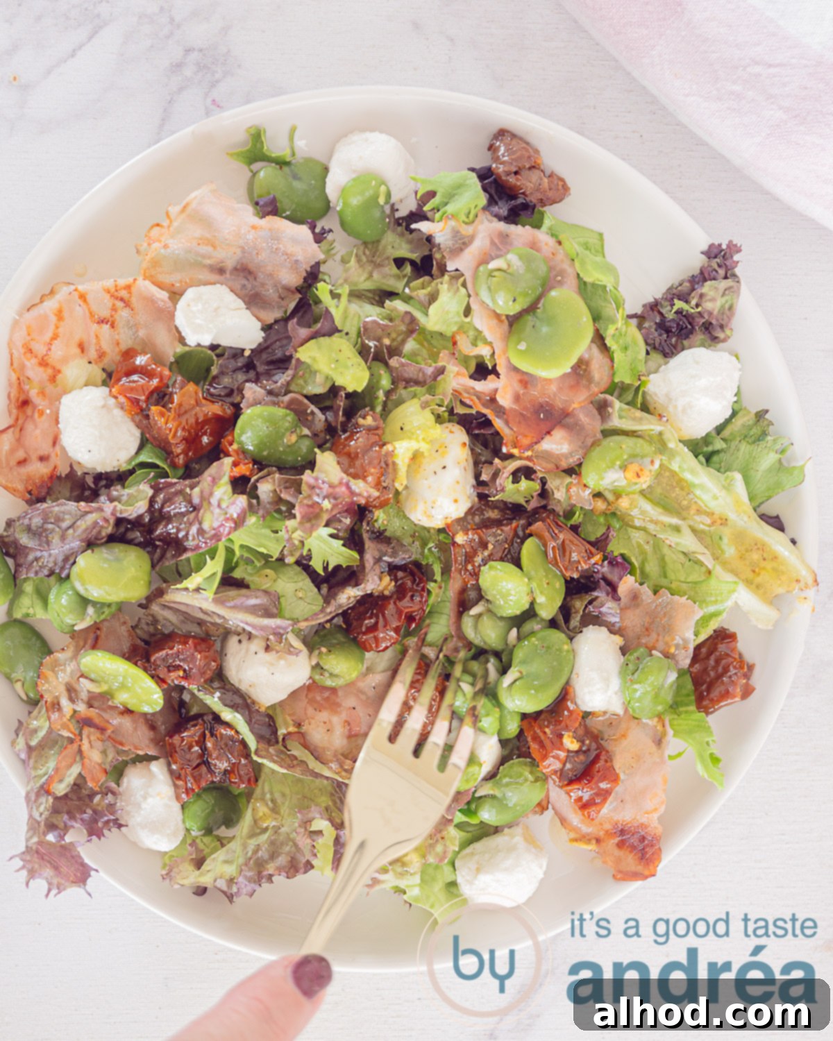 Spring Broad Bean, Bacon, and Goat Cheese Salad 2 A top-down view of a white plate showcasing a vibrant broad bean, bacon, and goat cheese salad. Mixed lettuce forms the base, topped with bright green fava beans, golden-brown bacon pieces, deep red sun-dried tomatoes, and crumbled white goat cheese. A fork is playfully positioned, piercing the fresh salad. The plate rests on a clean white background.