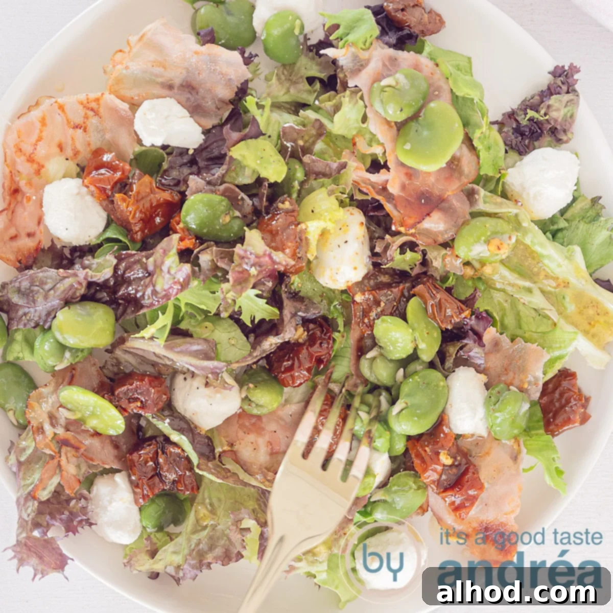 Spring Broad Bean, Bacon, and Goat Cheese Salad 6 A square photo with a round white plate with mixed lettuce, sun-dried tomatoes, bacon, broad beans and goat's cheese. A golden fork lies on it.