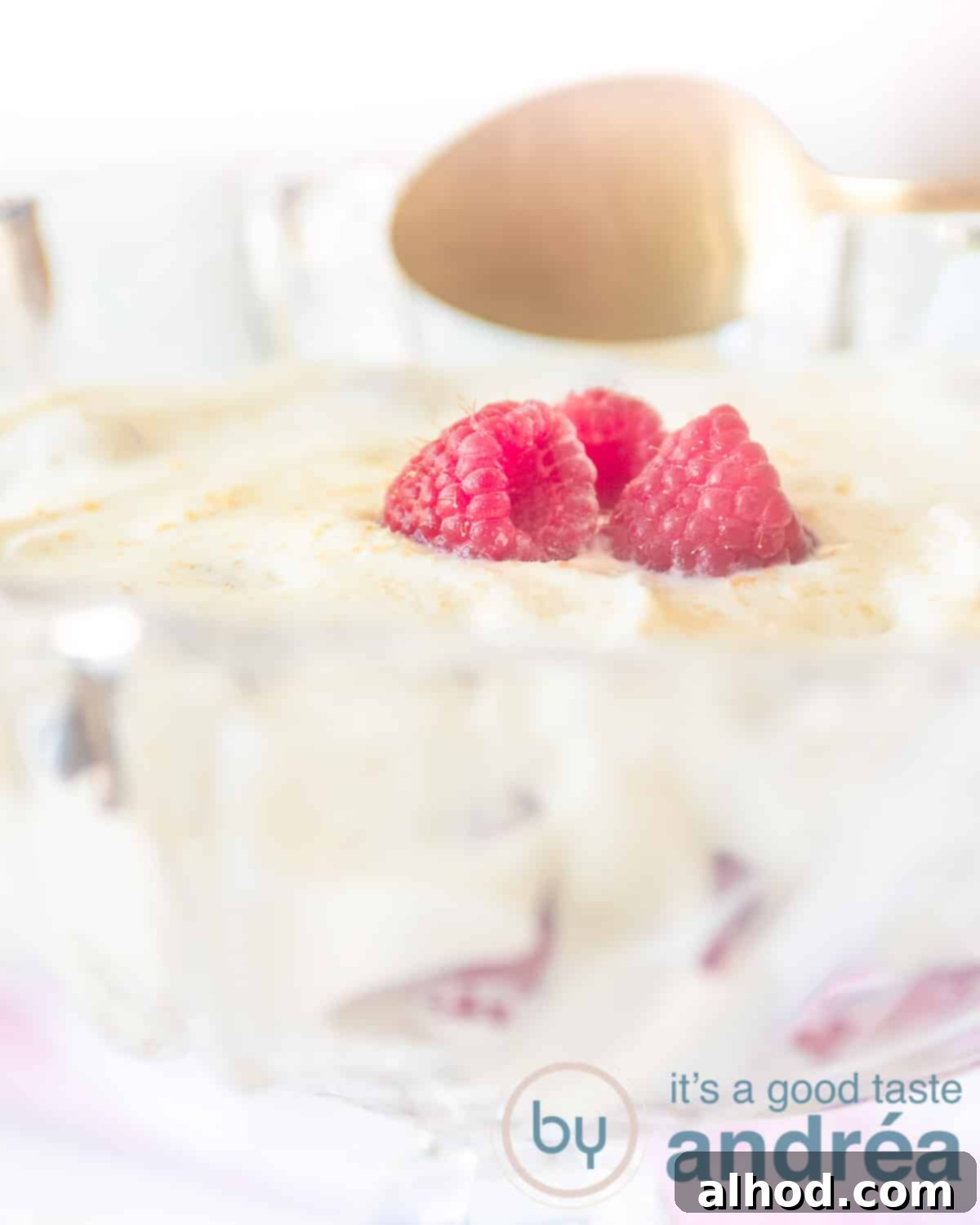 A glass bowl filled with raspberries, lady fingers and whipped cream. Topped with fresh raspberries. A golden spoon wants to take a bite