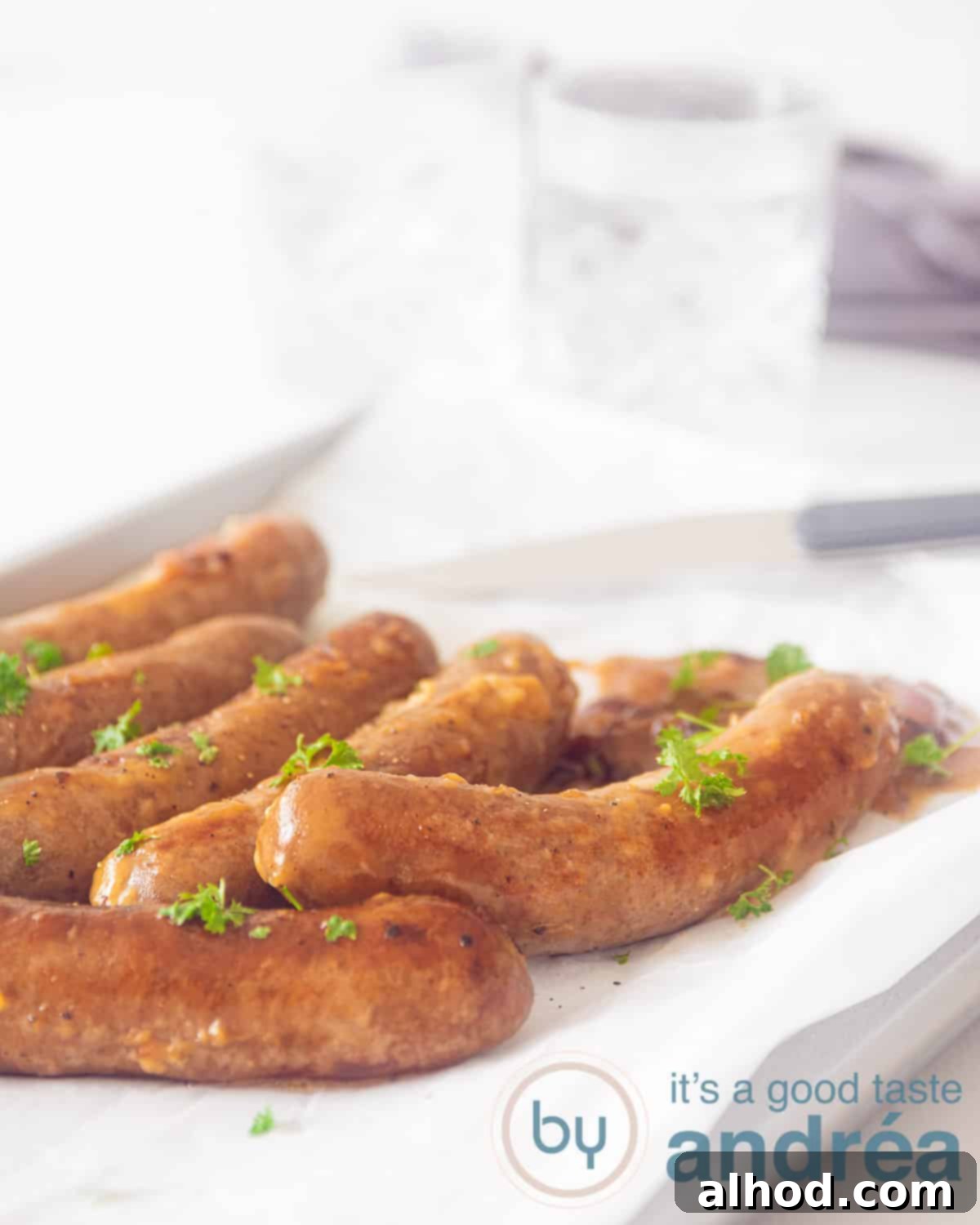 Beer-Braised Brats with Caramelized Onions 2 Sausage on a white baking paper garnished with parsley. A knife in the background