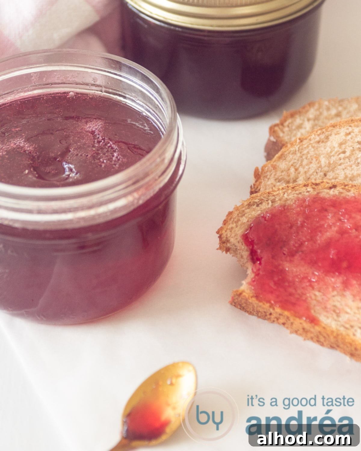 Rich Redcurrant and Port Jelly 2 An elevation photo showing part of a glass jar, open, containing vibrant redcurrant and Port jelly. On the right, two slices of bread are displayed, with the bottom slice generously smeared with the homemade jam. A golden spoon with a small amount of jelly rests at the bottom, inviting a taste.