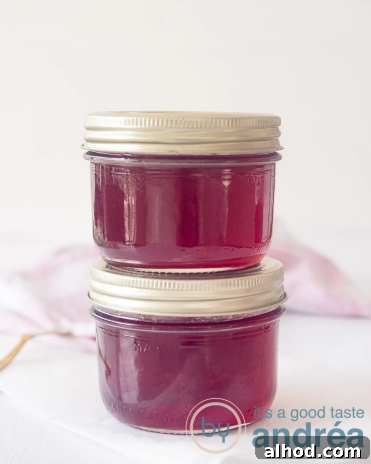 Rich Redcurrant and Port Jelly 5 Two neatly stacked glass jars of homemade redcurrant and Port jelly, each with a golden lid. They are presented against a clean white background, with a soft pink and white tea towel subtly visible in the background.
