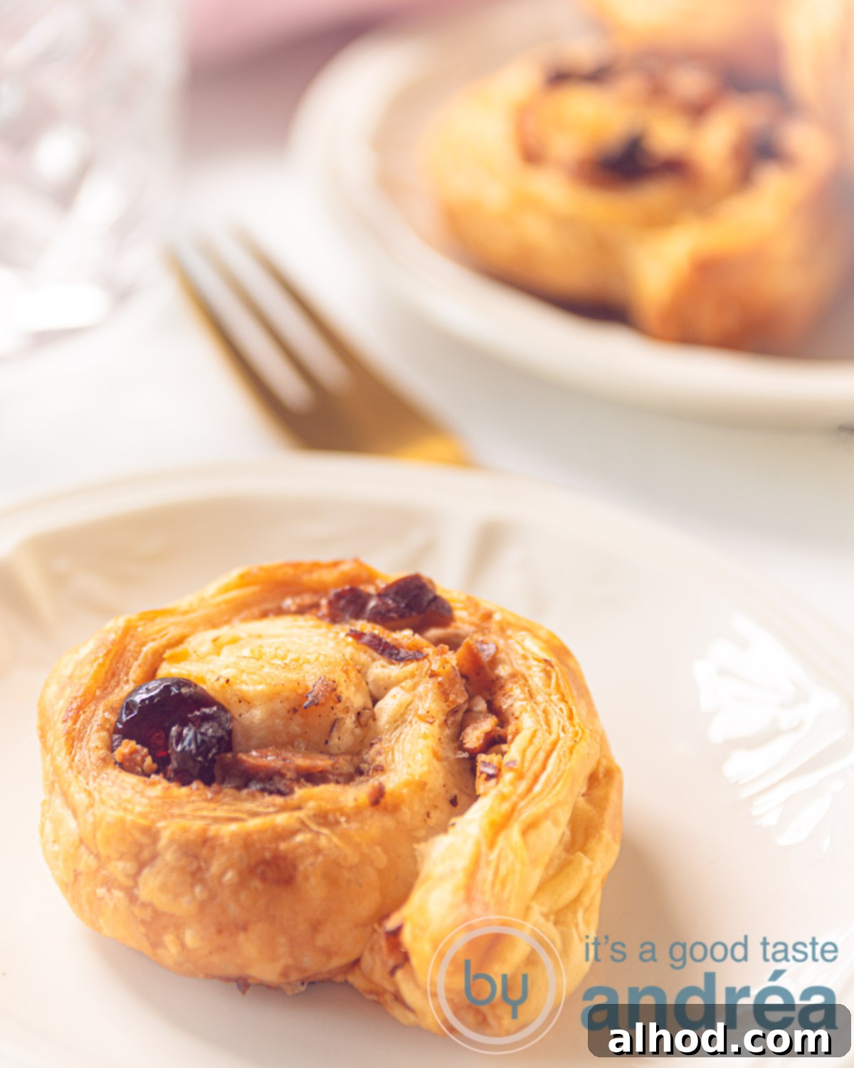 A white plate with a puff pastry cinnamon roll. A golden fork in the background as well as a plate with more sandwiches.