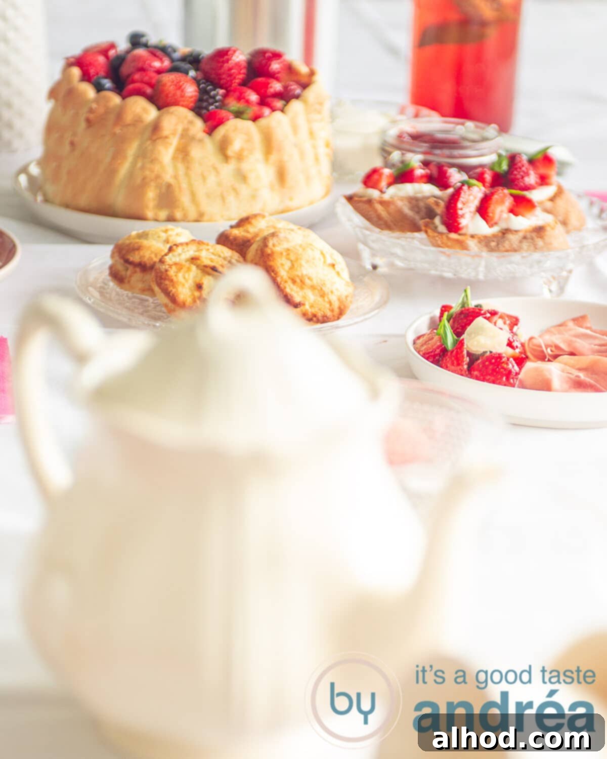 a white teapot in front and in the back high tea dishes like a Charlotte with fruit, bruschetta and strawberry lemonade