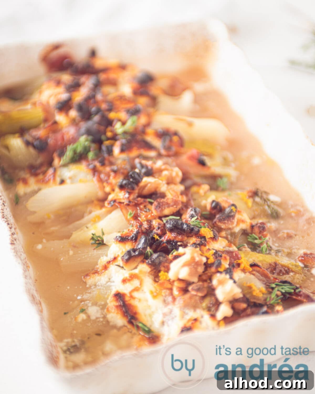 Belgian Endive Bake with Ham and Chevre 2 A beautifully presented white casserole dish filled with baked Belgian endive wrapped in Prosciutto, topped with melted goat cheese, scattered walnuts, and a drizzle of honey, ready to serve as a perfect side dish.