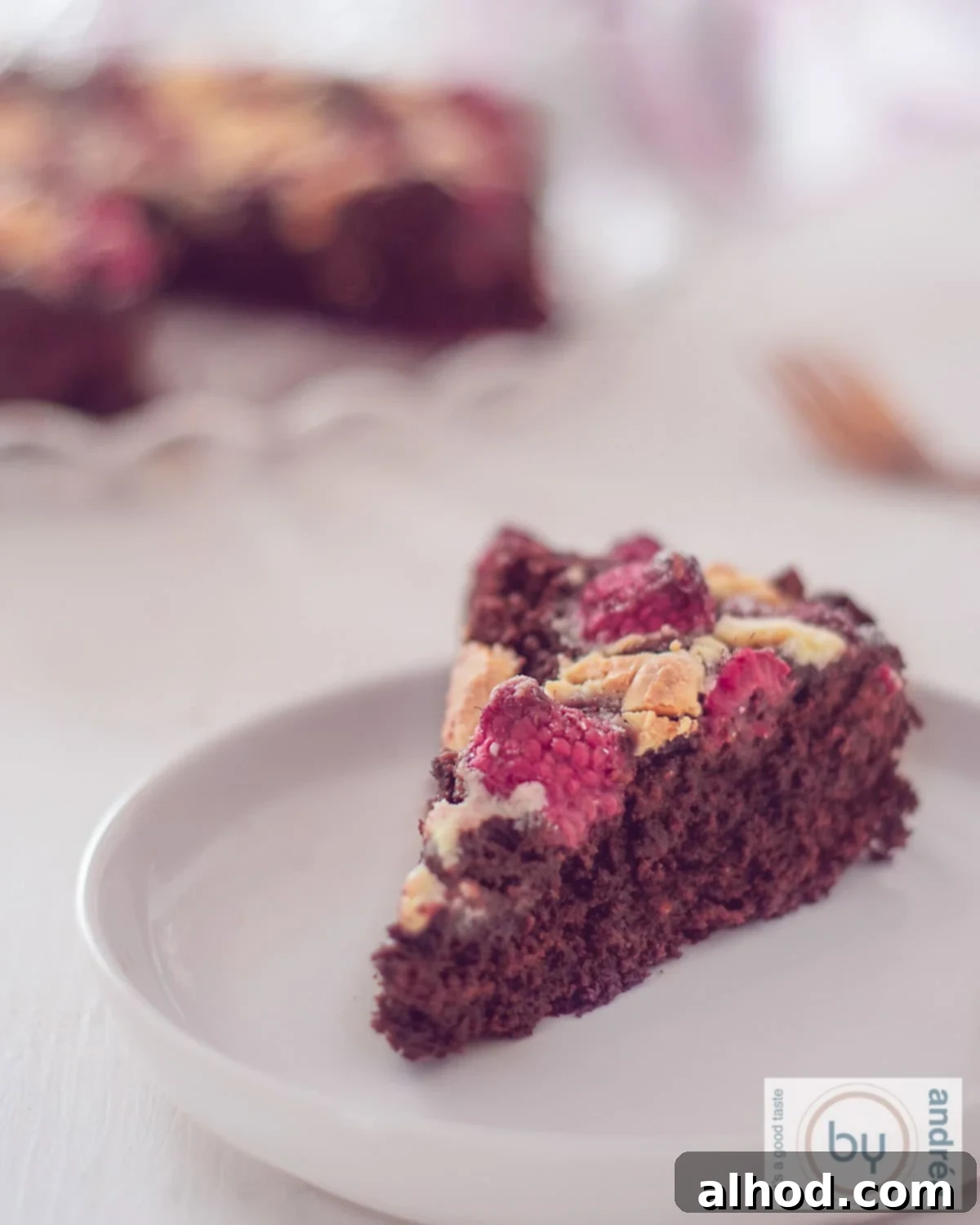 A slice of chocolate brownie with white chocolate chips and raspberries on a white plate. The rest of the cake is in the background.