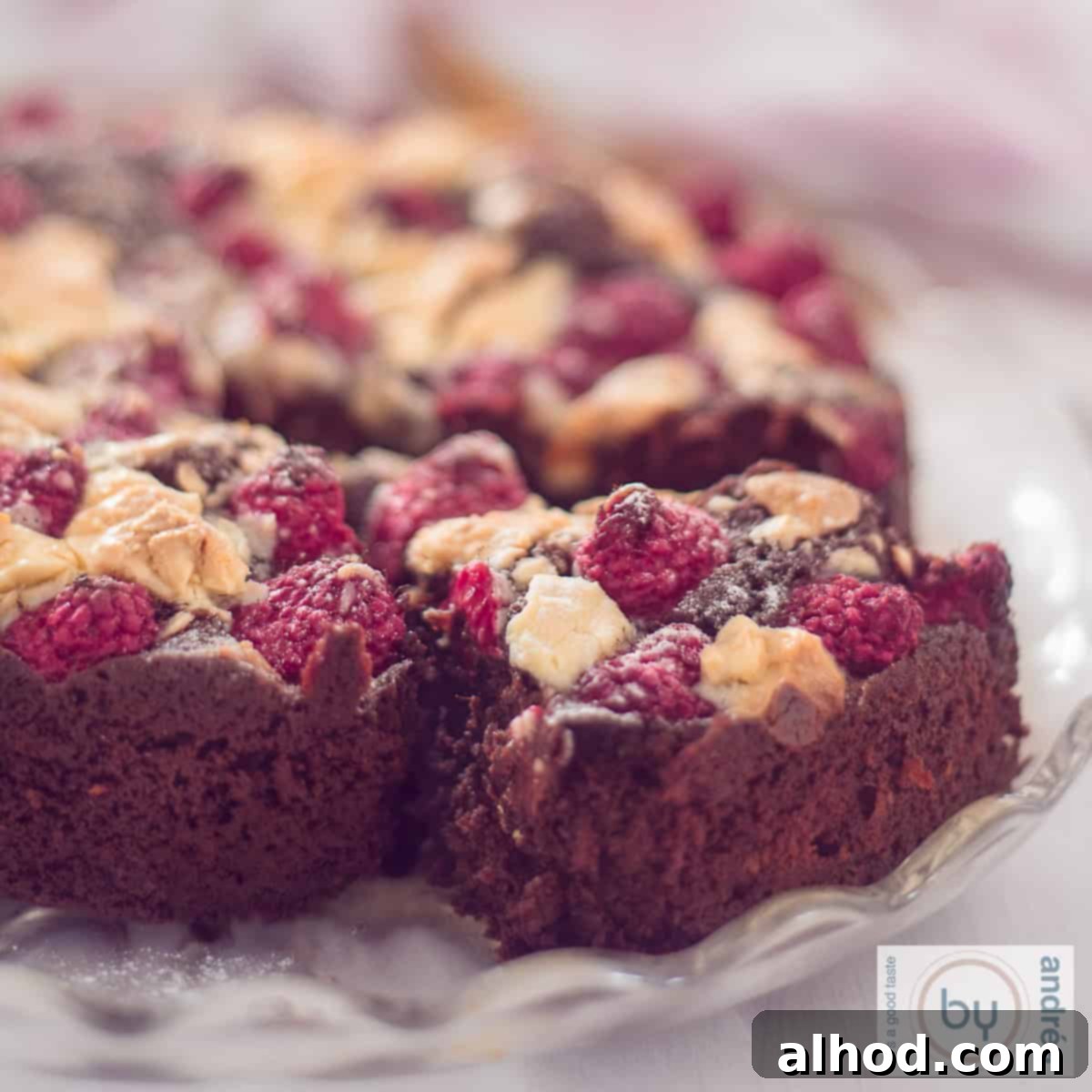A square photo of a brownie with a slice cut out of it. A topping of white chocolate and raspberries.