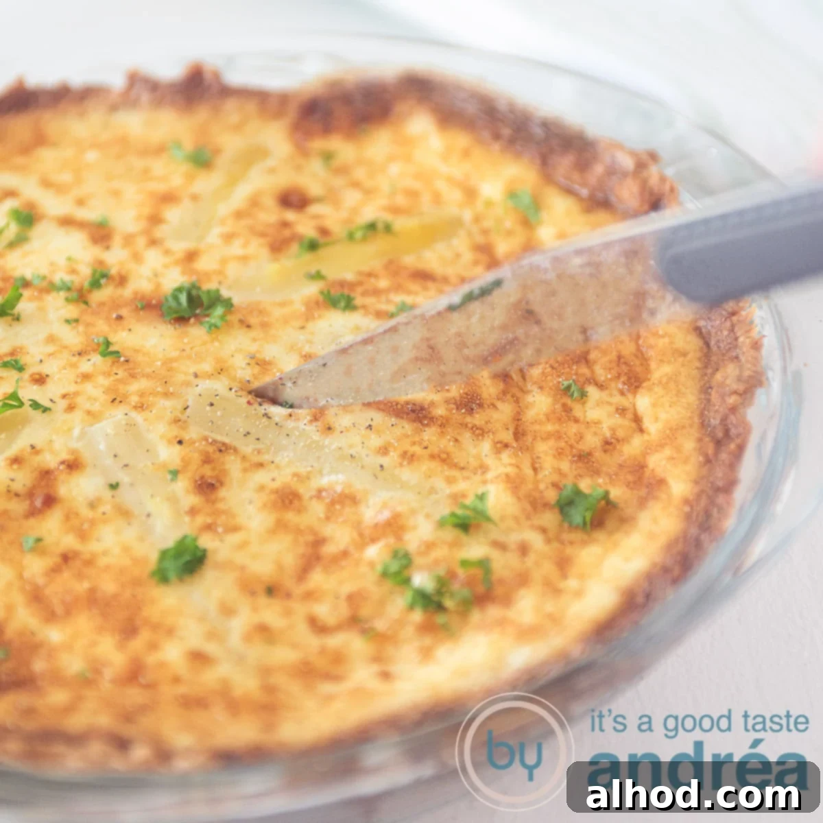 A square photo of a glass pie dish filled with a savory pie of asparagus, egg and cheeses. a knife cuts out a point