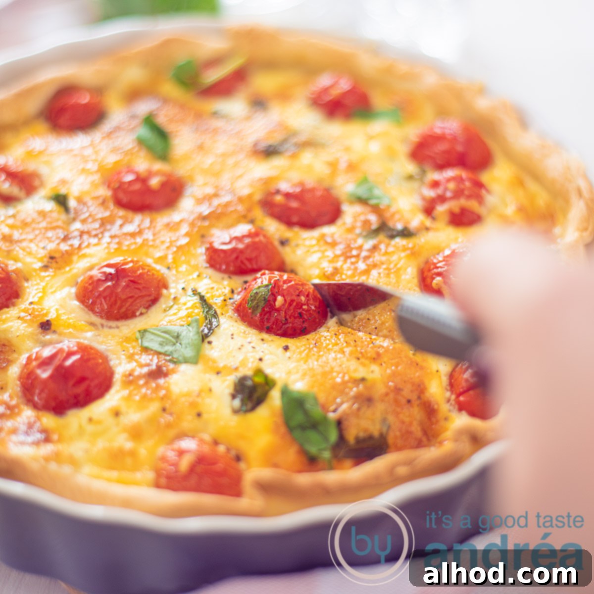 A Caprese Quiche in a grey pie dish, adorned with fresh tomatoes, mozzarella, and basil, with a wedge being cut out on a white surface.