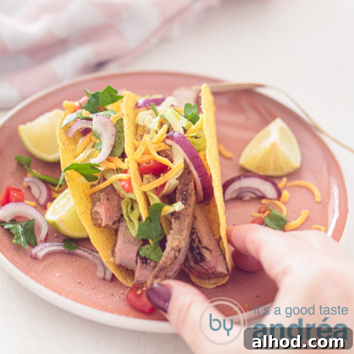 A vibrant pink plate showcasing grilled sirloin steak tacos filled with fresh vegetables and topped with cheddar cheese, with a hand holding one taco.