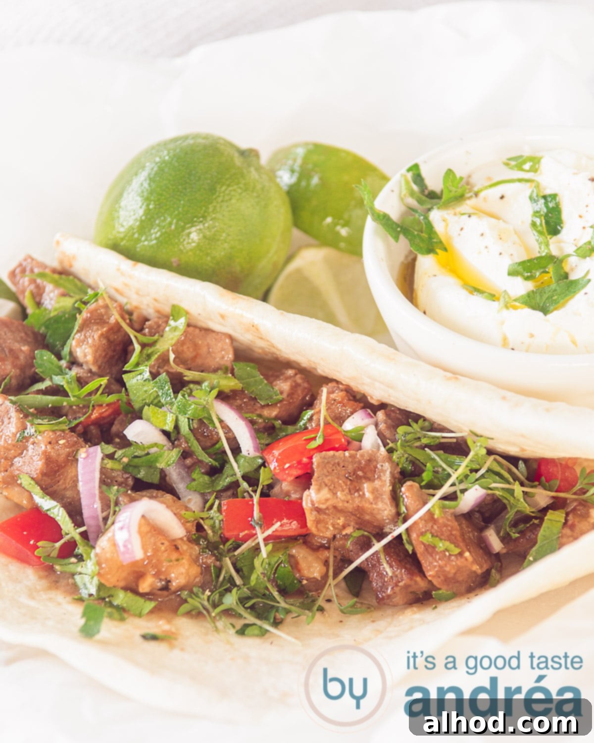 A white plate featuring a tortilla filled with tender Carne Guisada, garnished with fresh tomato and onion, with lime and crème fraîche in the background.