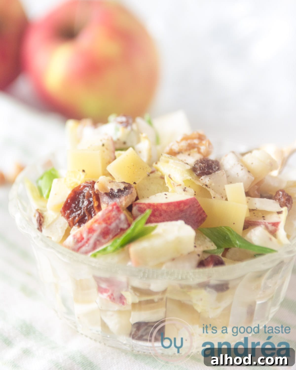 A full, inviting bowl of Brussels endive (chicory) salad, beautifully mixed with apple, raisins, and walnuts. Two fresh apples are positioned appealingly in the blurred background.