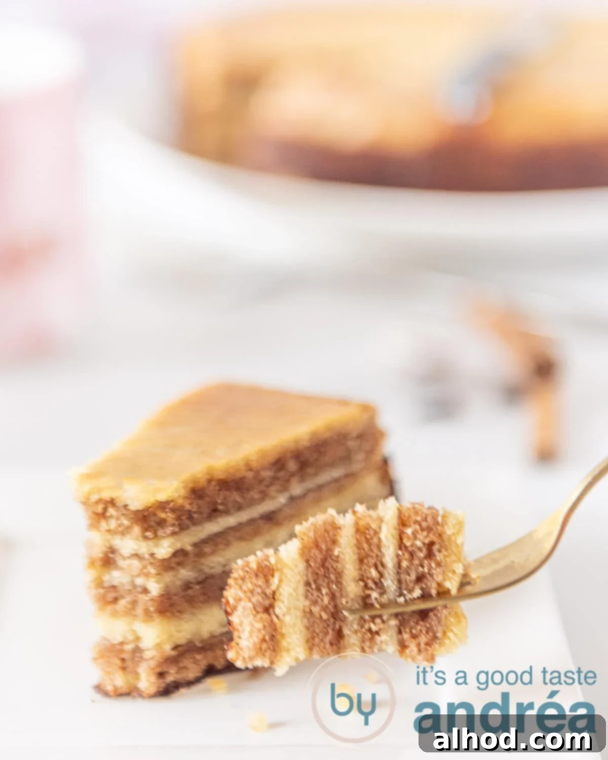 A fork holding a bite of the Indonesian Thousand Layers Cake (Lapis Legit). In the background, a whole cake and a slice are visible, emphasizing its delicate texture and the numerous layers.
