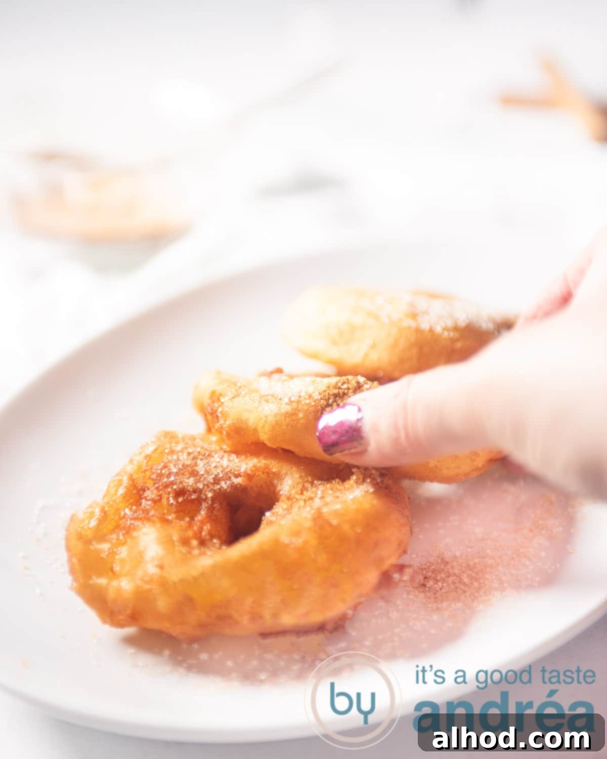 A hand taking an apple fritter from a white bowl with three fritters