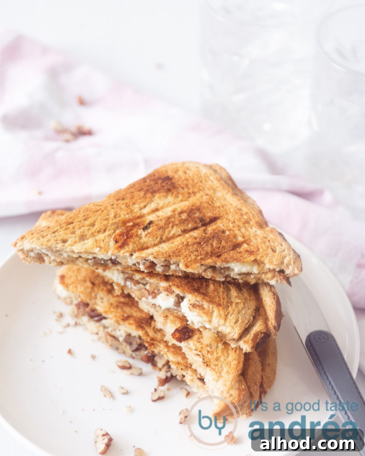 A white plate with three half sandwiches filled with cheese and nuts. A knife on the right. in the background a white background, a pink white checkered tea towel and two crystal glasses