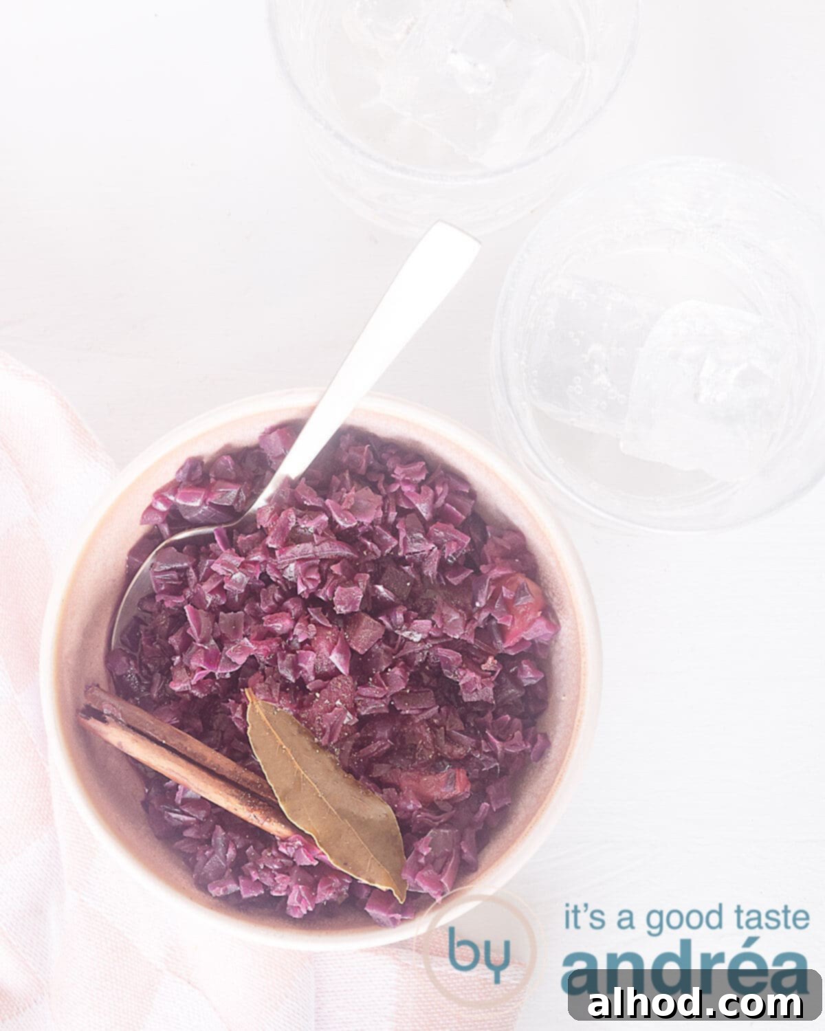 Sweet and Sour German Red Cabbage 2 A photo from above of a vibrant pink dish filled with sweet and sour red cabbage, garnished with bay leaf and cinnamon, set against a pristine white background. Two glasses are visible in the top right corner.