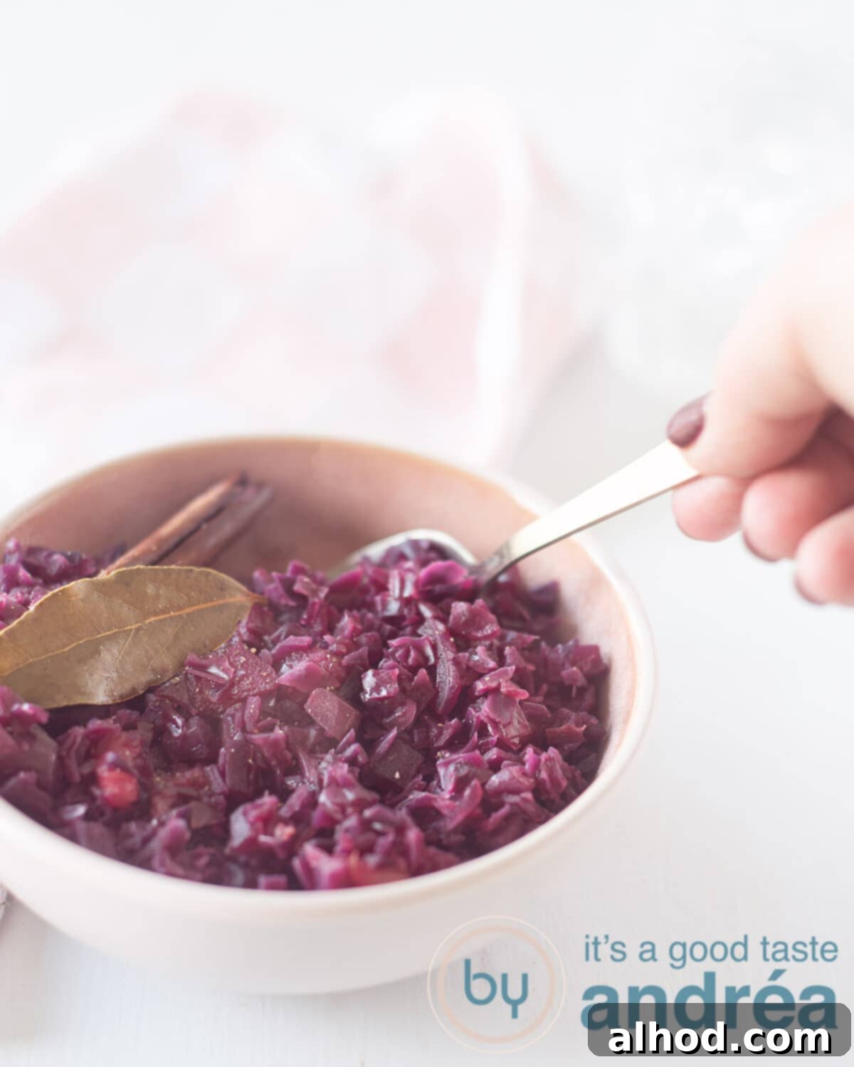 Sweet and Sour German Red Cabbage 5 A close-up photo of vibrant red cabbage in a pink serving dish, garnished with a bay leaf and cinnamon stick. A hand reaches in with a spoon, ready to serve a portion.