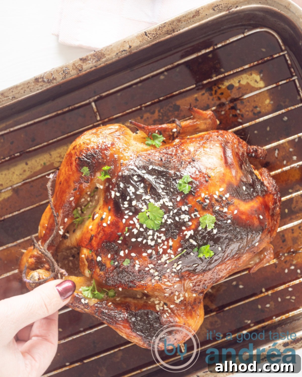 Easy Sticky Soy Sauce Chicken 2 A photo from above of a perfectly roasted whole soy sauce chicken on a wire rack in a baking pan, garnished with sesame seeds and parsley. A hand is reaching for a chicken leg.