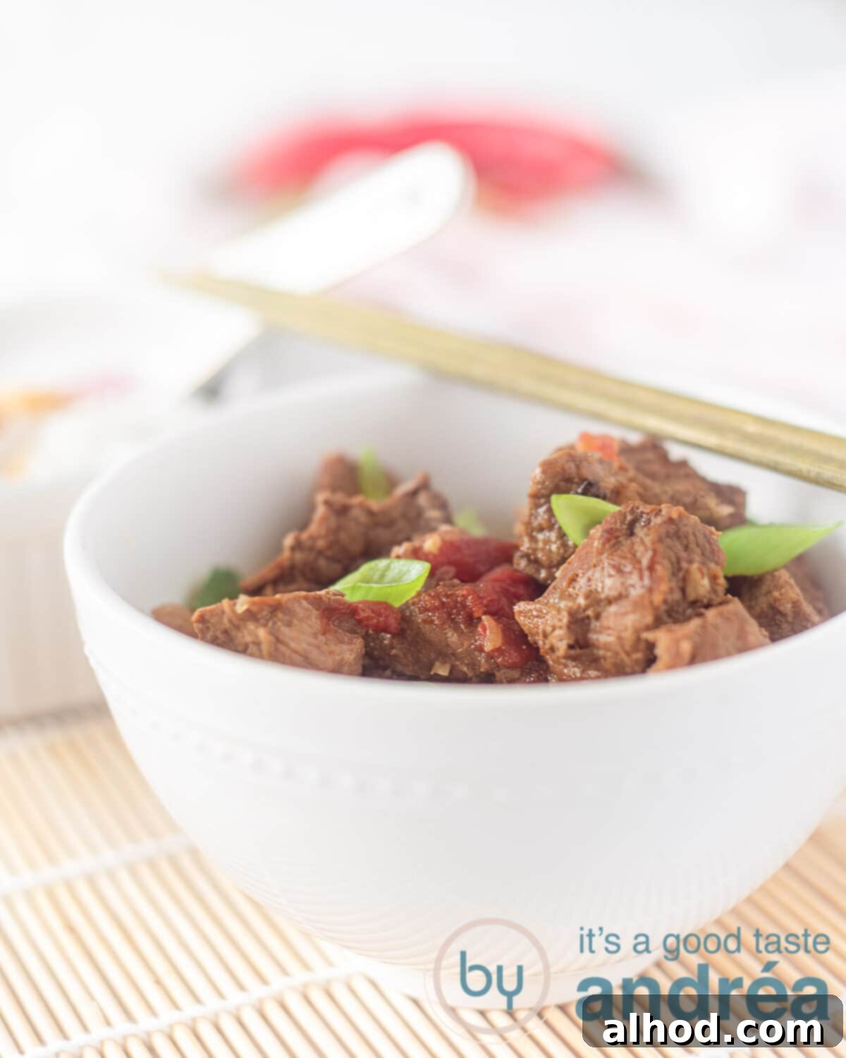 Sweet & Savory Slow-Simmered Beef 2 A white bowl filled with slow-cooked Semur Daging, garnished with fresh chilies and vibrant spring onions. Two wooden chopsticks rest on the side of the bowl, which is placed on a rustic wicker mat.