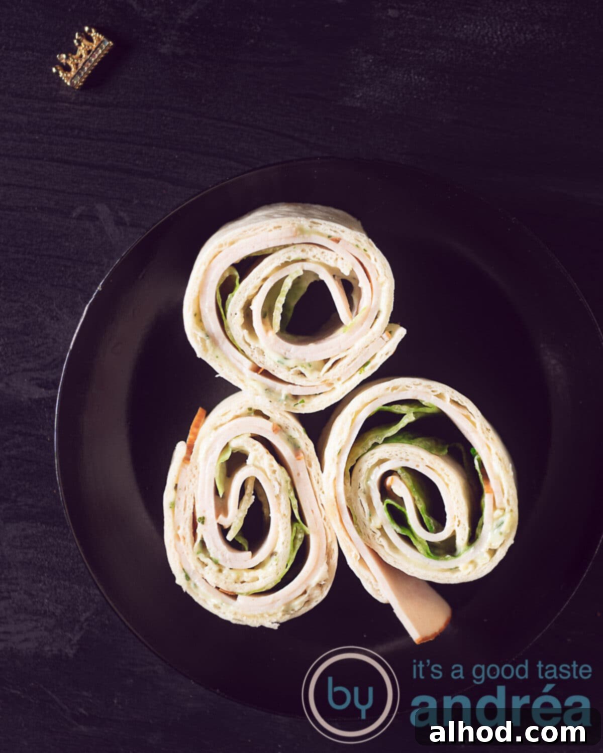 Creamy Chicken Tortilla Pinwheels 2 A black plate with three rolled tortilla rolls featuring chicken breast, crisp iceberg lettuce, and creamy herb cheese, arranged on a dark board against a black background. This image showcases a tasty snack perfect for any party: cold wraps with chicken and herb cream cheese.