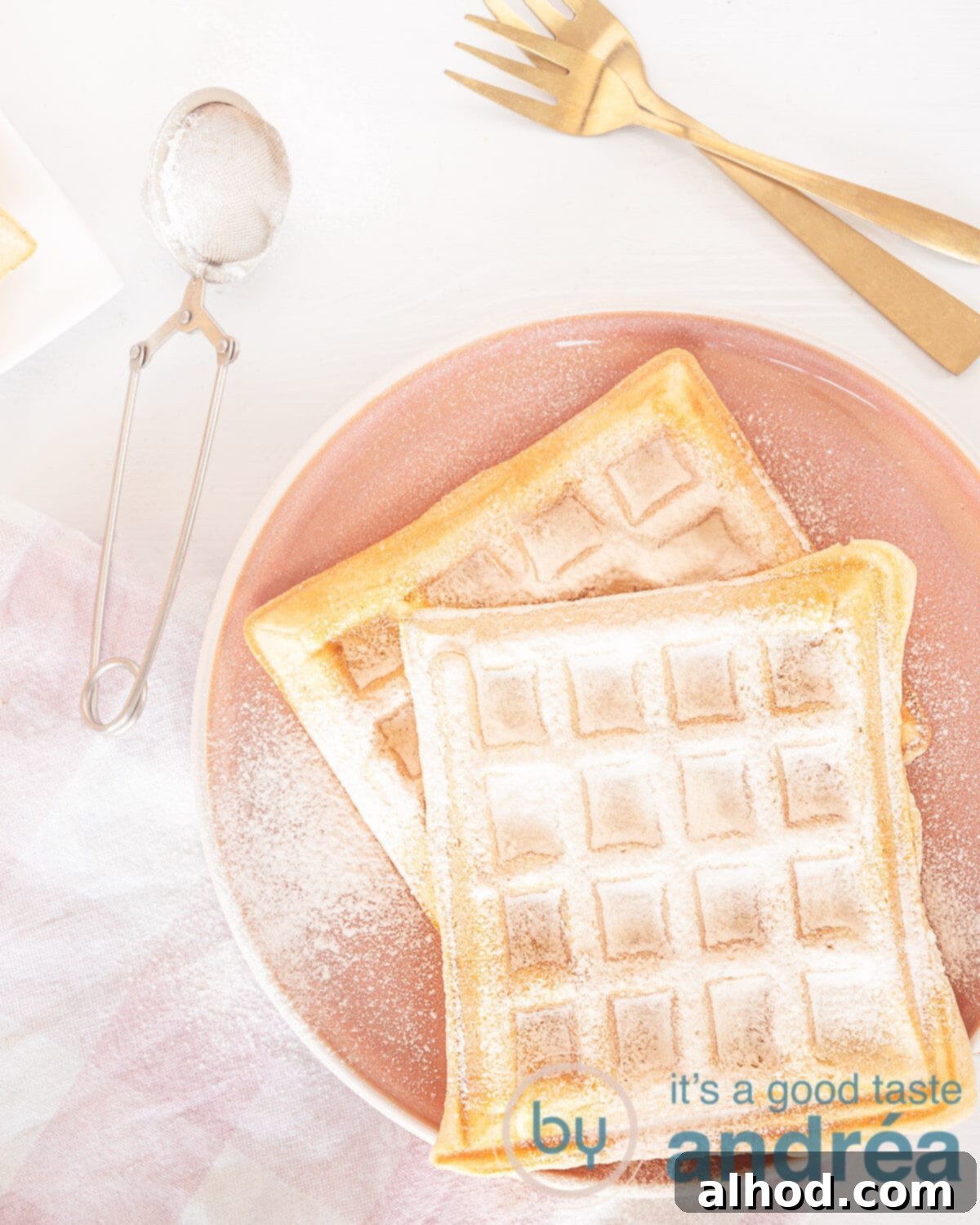 A photo from above of old fashioned Dutch waffles on a pink plate, dusted with icing sugar. In the lower left corner a pink white tea towel. A sieve with powdered sugar and two golden forks. A white background, inviting the viewer to enjoy.