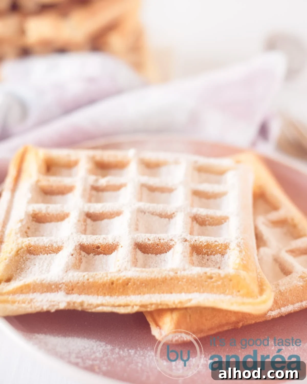 Authentic Dutch Wafels 6 A pink plate with two waffles sprinkled with powdered sugar. in the background a pink and white checkered tea towel, showcasing a delightful homemade Dutch waffle.