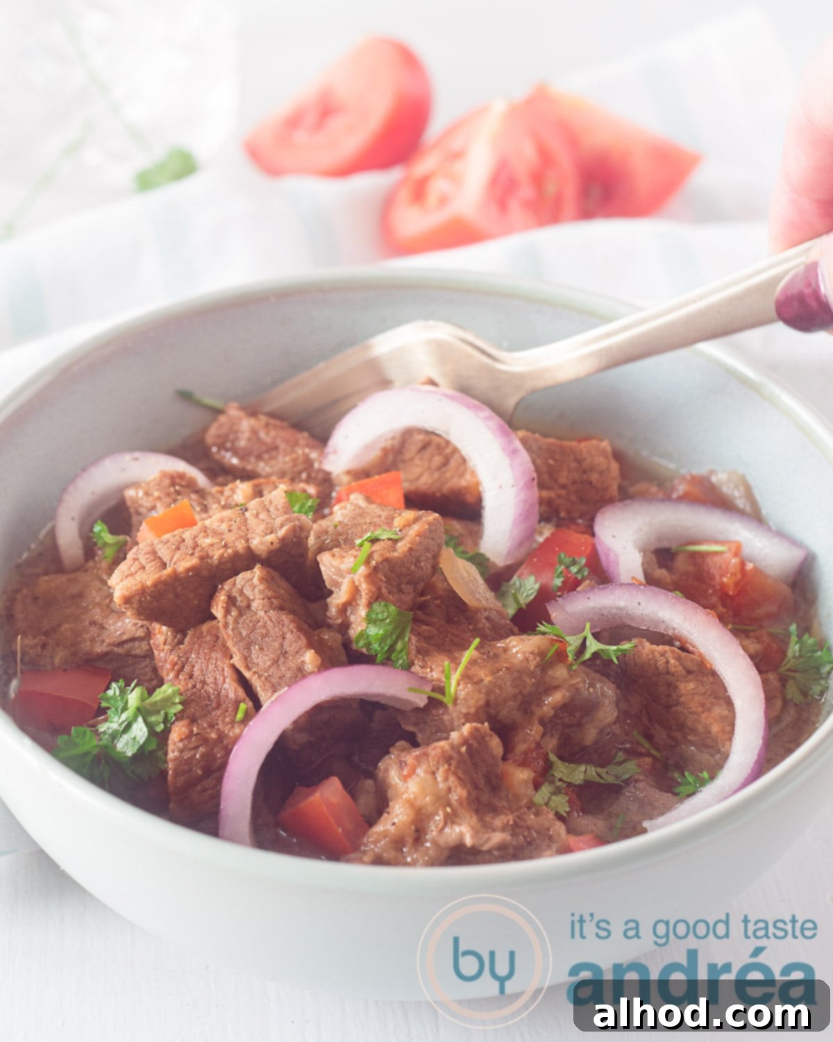 Hearty Slow Cooker Mexican Beef Stew 2 A blue bowl filled with Tex Mex Carne Guisada, garnished with onion and tomato. A golden fork spears a piece of tender beef. Fresh tomatoes in the background provide a vibrant touch.
