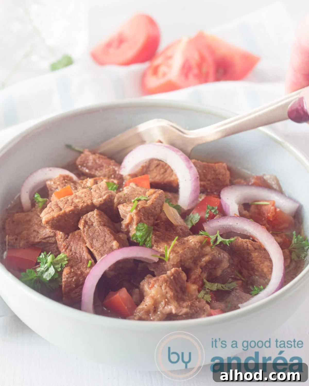 Savory Slow Cooker Mexican Carne Guisada 2 A blue bowl filled with Tex Mex Carne Guisada, garnished with onion rings and a slice of tomato. A golden fork spears a piece of tender beef. Fresh tomatoes are visible in the soft background.