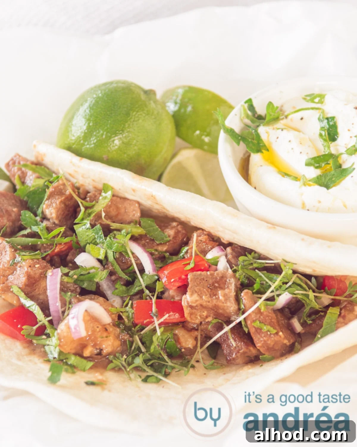 Savory Slow Cooker Mexican Carne Guisada 6 A white plate with a tortilla filled with Carne Guisada, tomato and onion. Lime and a bowl of crème frache in the background
