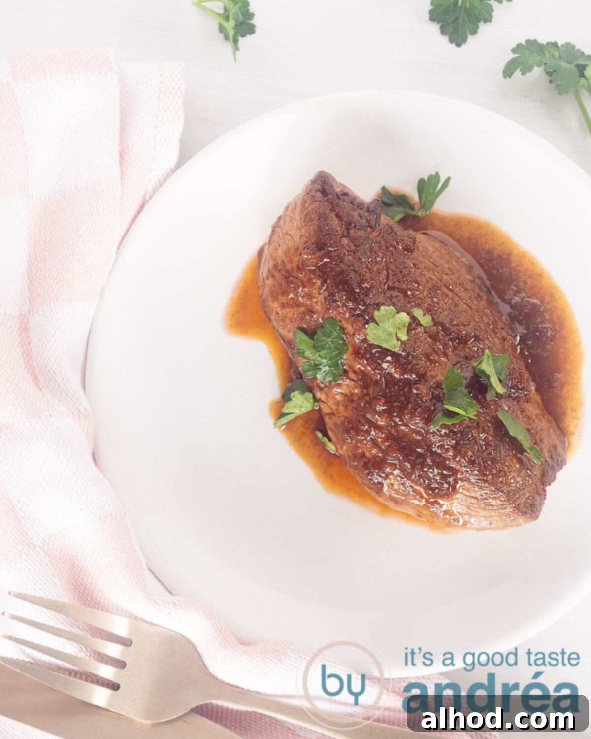 A photo from above of a perfectly seared, tender Bali steak generously coated in its signature brown gravy, garnished with fresh parsley on a pristine white plate, set against a clean white background. The steak looks incredibly juicy and inviting.