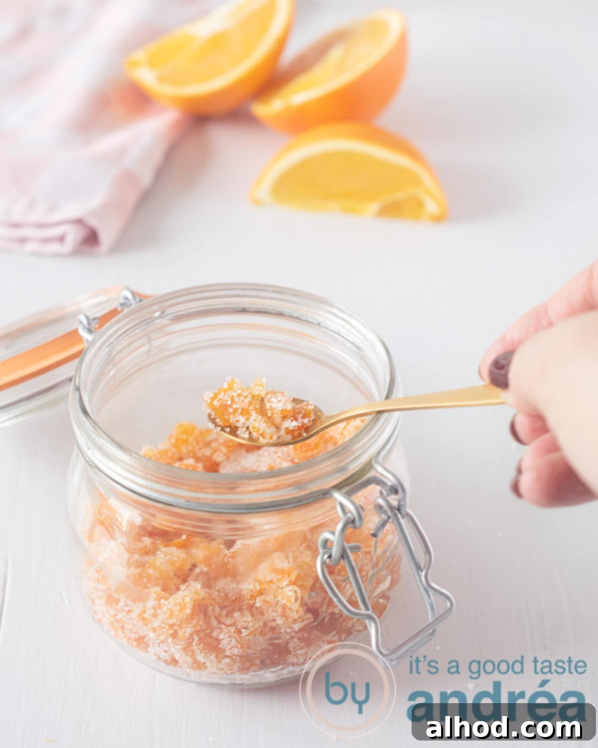 A white background with a jar of glistening candied orange peel in the foreground. A golden spoon is shown taking a scoop of the sweet treat. In the background, a charming pink and white checkered tea towel and fresh orange wedges complete the inviting scene.
