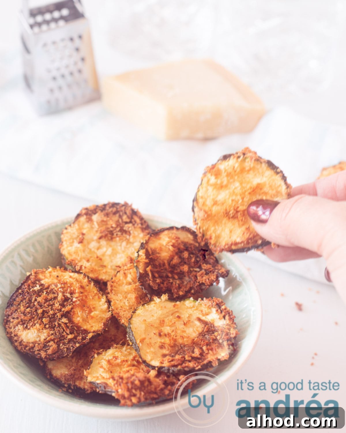 Crispy Air Fryer Zucchini Chips with Parmesan 5 A hand reaching for a golden-brown Air Fryer Parmesan Zucchini Chip from a full bowl. The background shows Parmesan cheese and a grater, emphasizing the fresh ingredients.