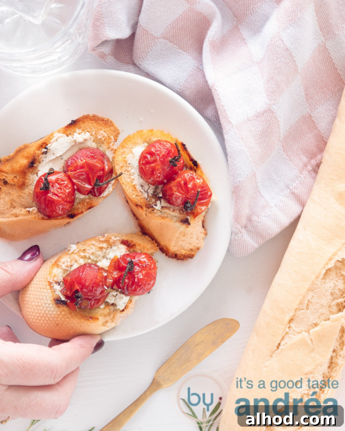 Creamy Goat Cheese and Roasted Tomato Bruschetta 2 A top-down view of a white plate holding three slices of bruschetta with goat cheese and stewed tomatoes. A hand reaches for one slice. In the background, a baguette and a white and pink tea towel.