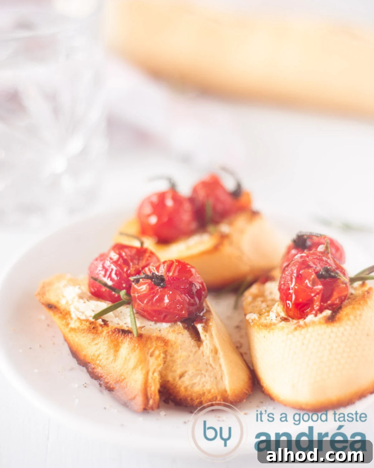 Creamy Goat Cheese and Roasted Tomato Bruschetta 5 An elevation photo showing three grilled baguette slices topped with creamy goat cheese and stewed cherry tomatoes, arranged on a white background. A whole baguette and a glass are blurred in the background.