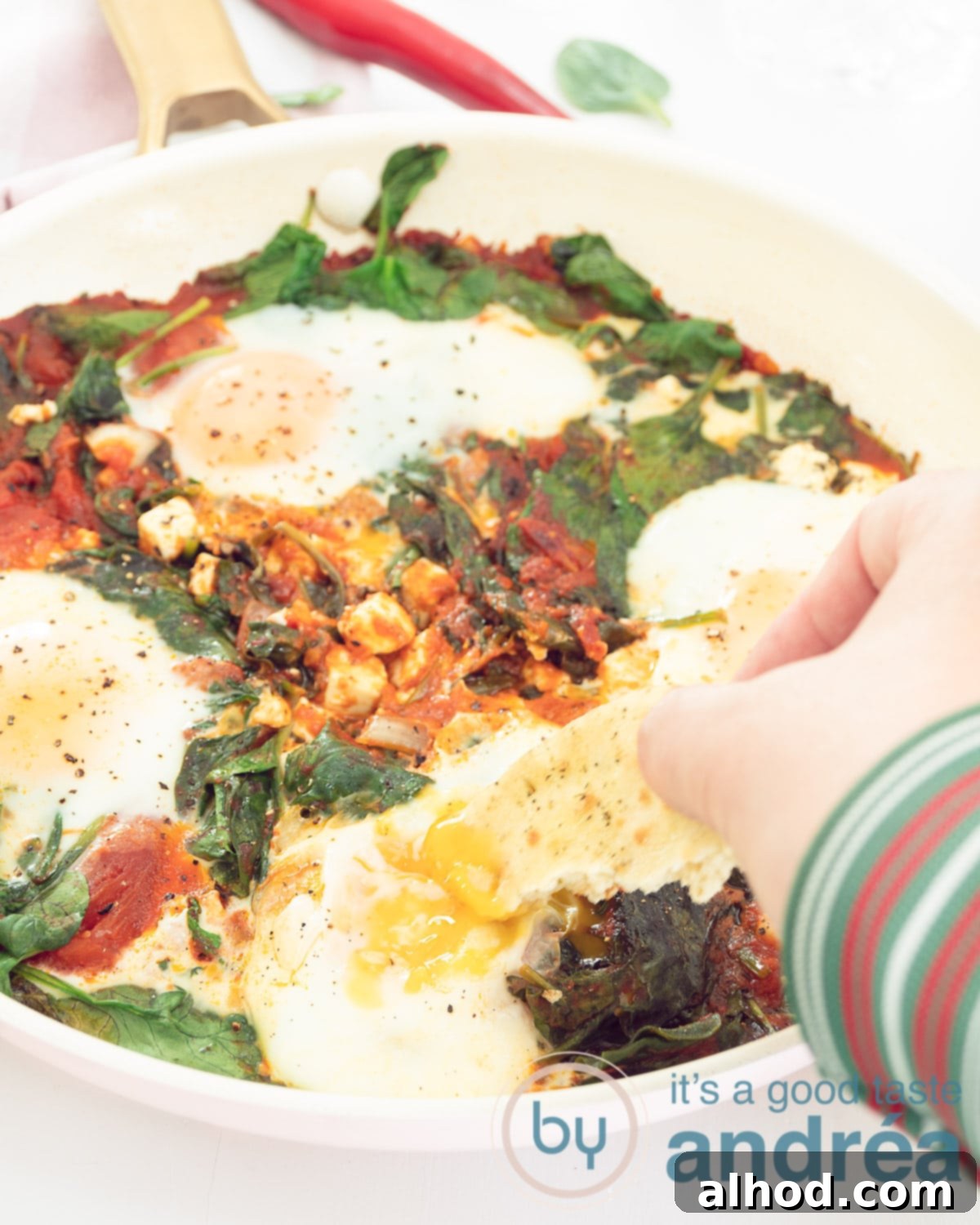 A pink pan filled with shakshuka with egg, feta and spinach. A hand dips some bread.