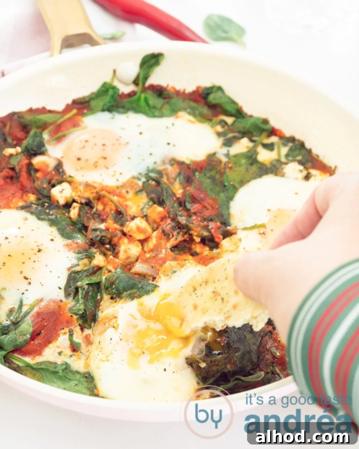 Effortless Spinach Feta Shakshuka with Eggs 2 A pink pan filled with shakshuka with egg, feta and spinach. A hand dips some bread.