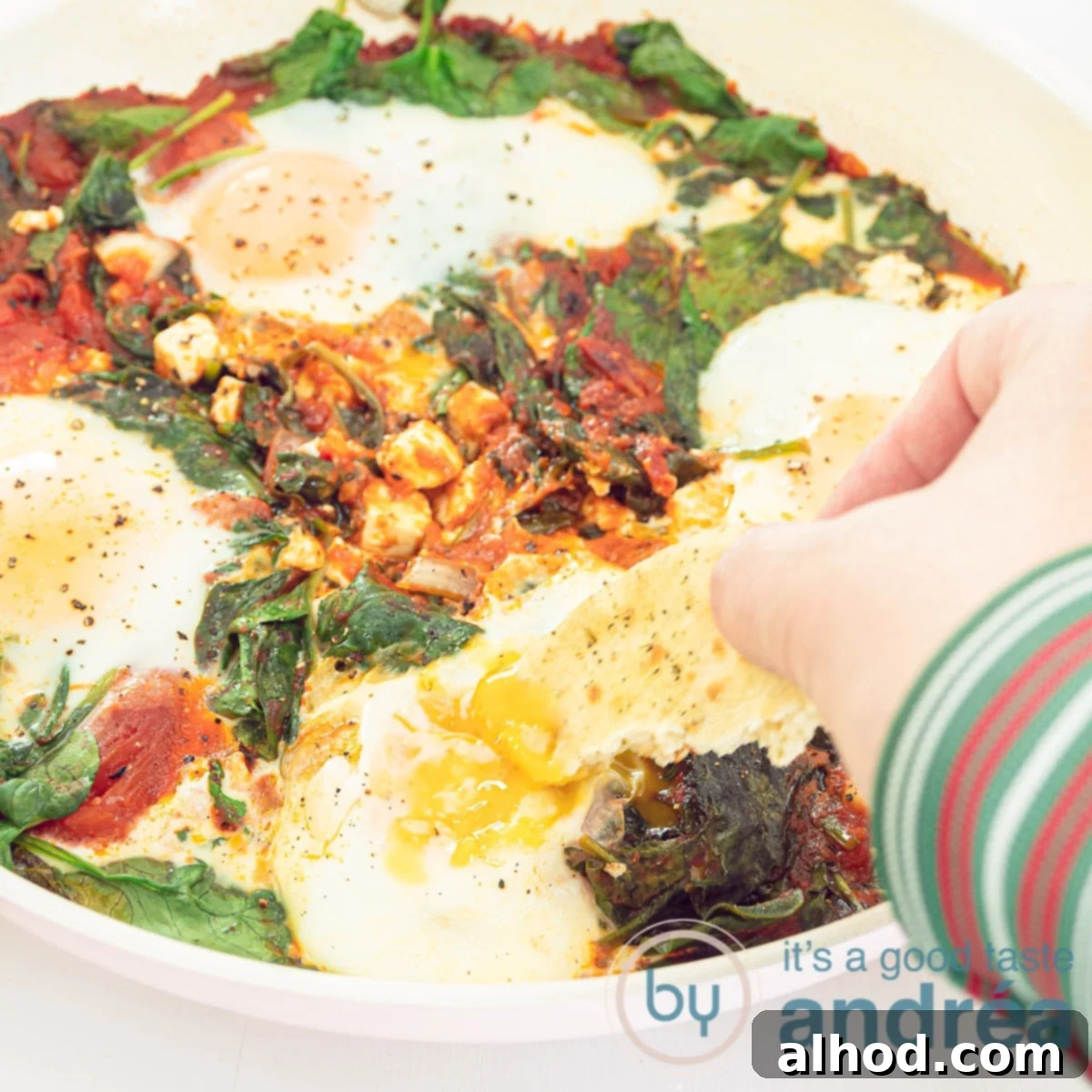 Effortless Spinach Feta Shakshuka with Eggs 4 A square photo with shakshuka in a frying pan. A hand dips into the poached egg with some bread.