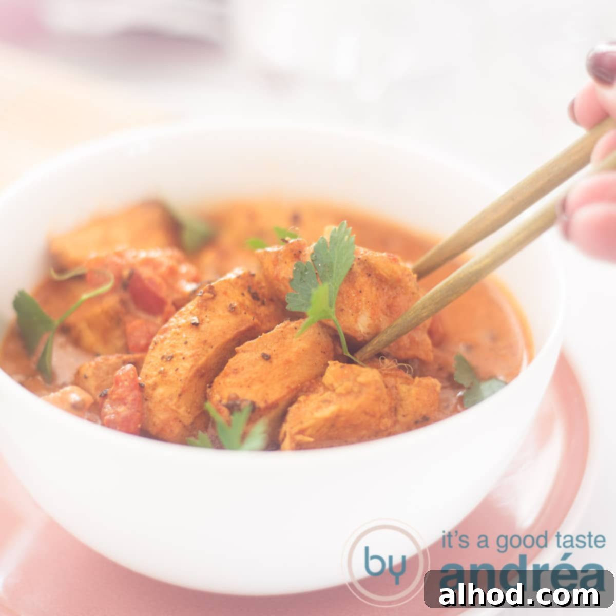 Effortless Slow Cooker Butter Chicken 6 A square shot showing a white bowl on a pink plate filled with slow cooker butter chicken. Two chopsticks grab a piece of chicken, highlighting its tender texture.