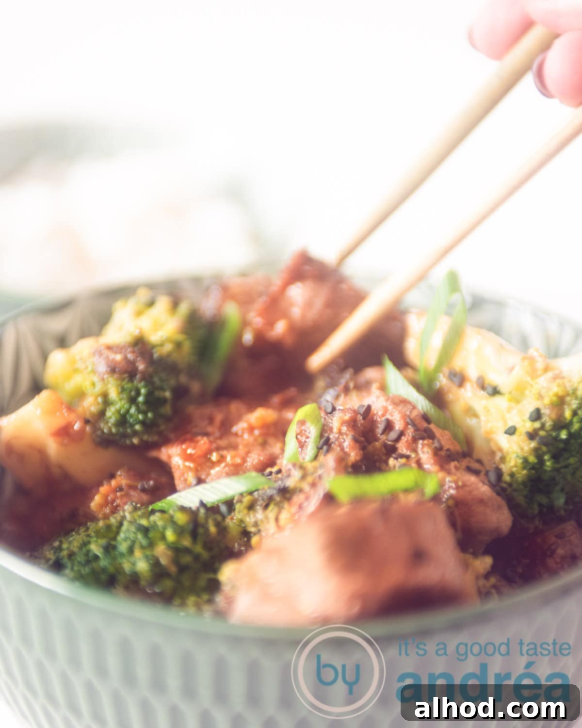 Effortless 20-Minute Teriyaki Beef and Broccoli Dinner 2 A bowl filled with steak, broccoli florets, mixed with a teriyaki sauce on a white background. Two chopsticks take a diced steak from the bowl.