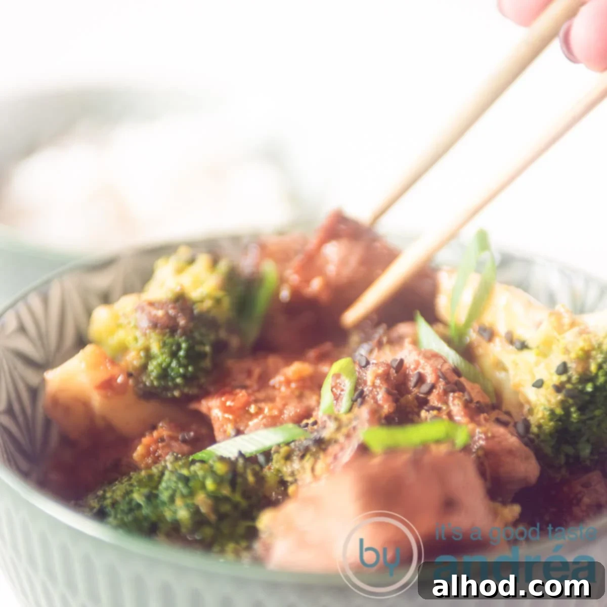 Effortless 20-Minute Teriyaki Beef and Broccoli Dinner 6 A square photo where two chopsticks take a cube of steak from a green bowl filled with broccoli, steak and sauce.
