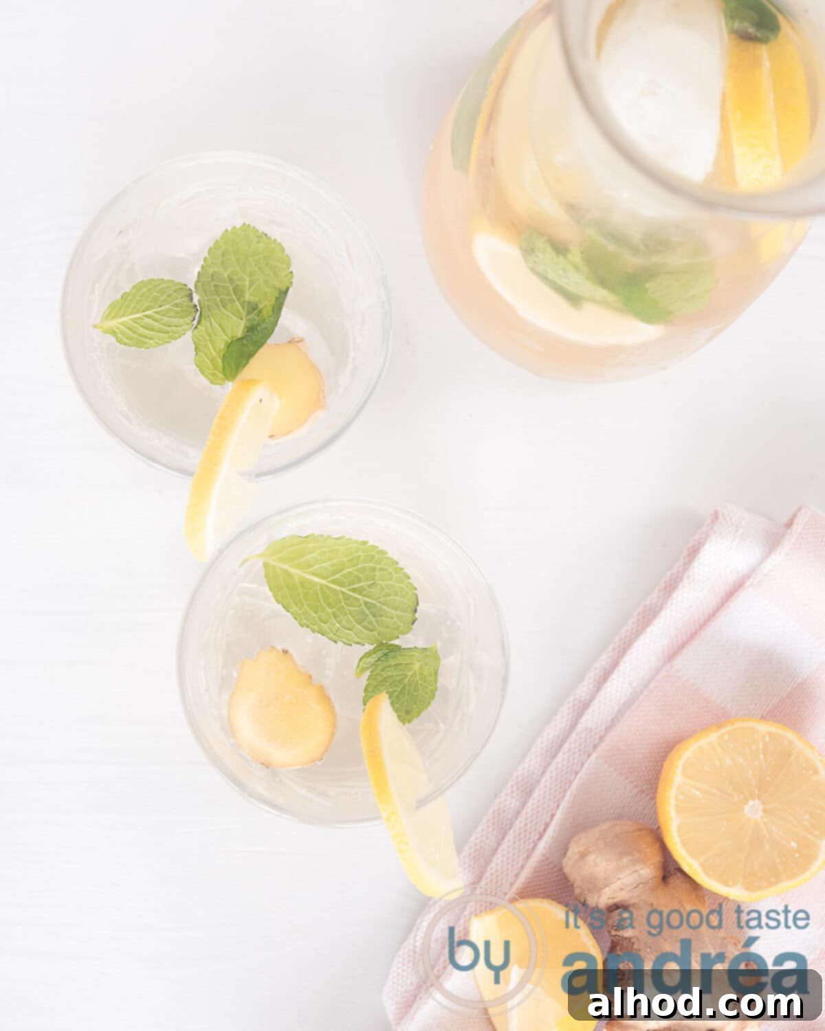 A photo from above with two glasses filled with lemon-ginger lemonade and a carafe. At the bottom a pink white tea towel with lemon and a press