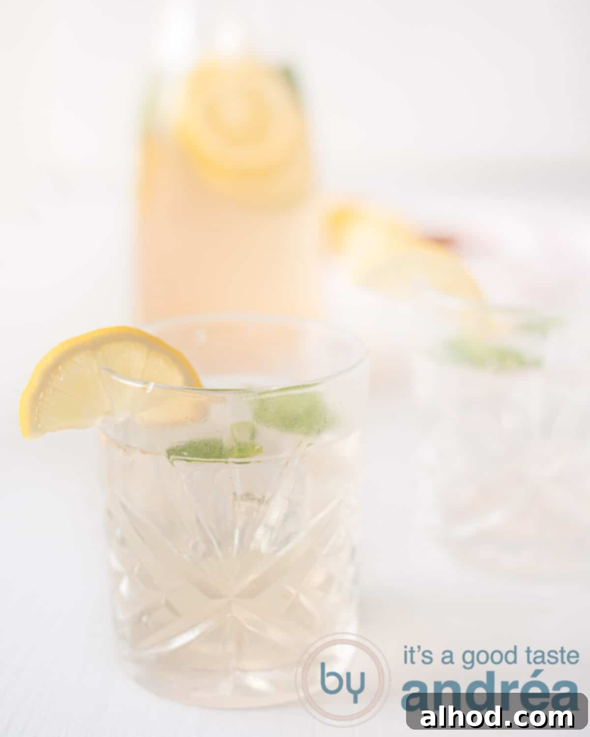An elevation photo with a glass filled with lemonade with ginger and lemon. A carafe in the background everything on a white background