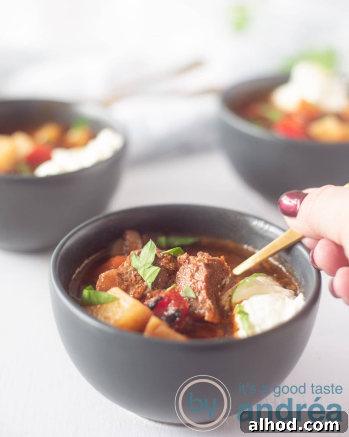 Hearty Slow Cooker Beef Goulash Soup 2 A hand scooping a spoonful of goulash soup from a gray bowl. You can see two other bowls in the background.