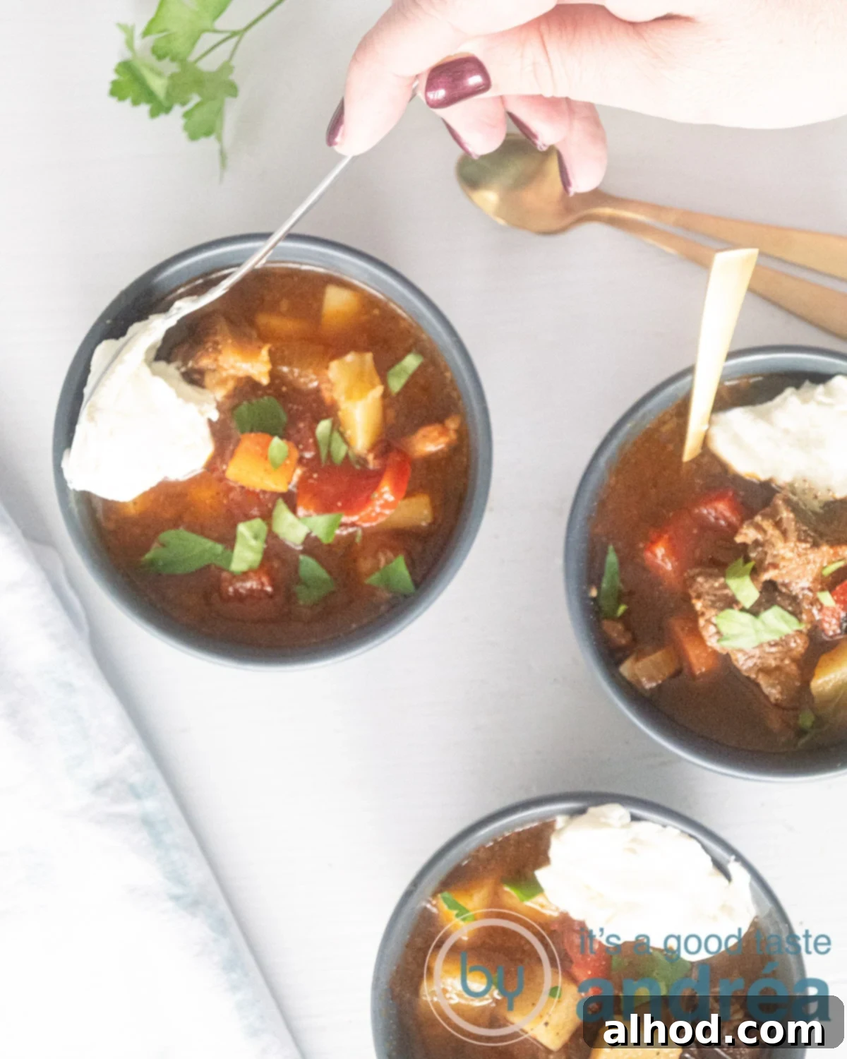 Hearty Slow Cooker Beef Goulash Soup 5 A hand scooping a spoonful of crème frache into the soup. Two more soup bowls are in the picture. All on a white background