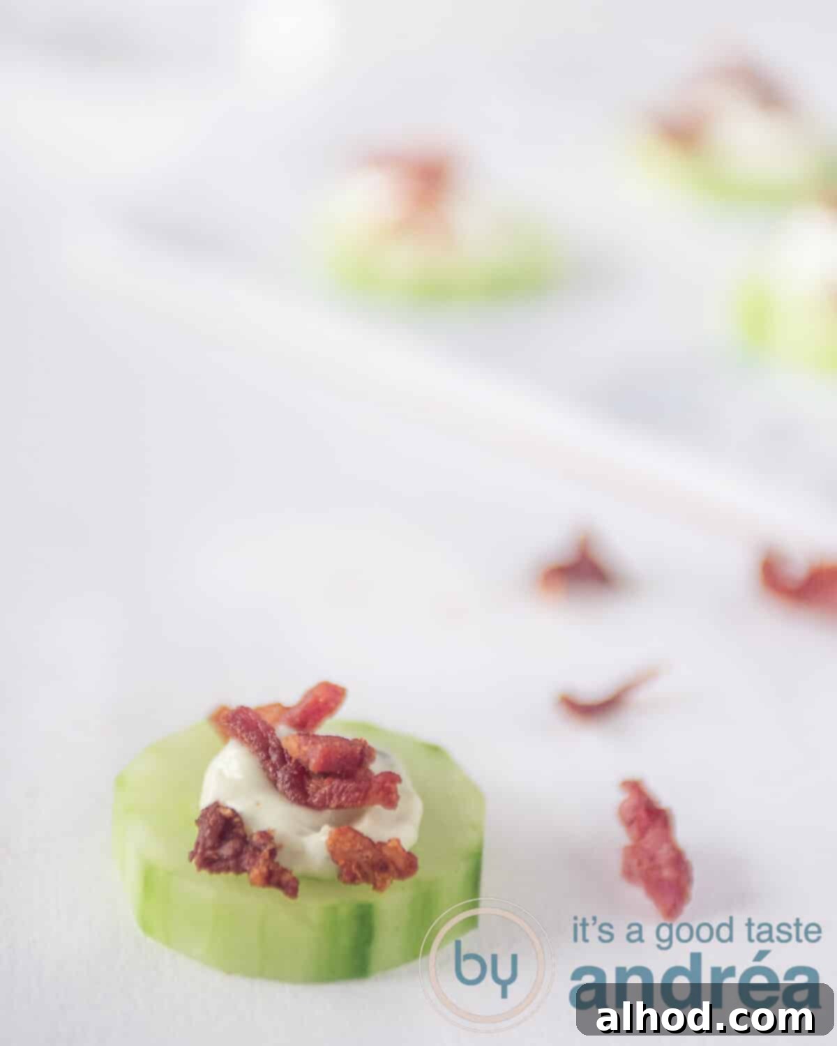 Leek Dip Cucumber Bites with Crispy Bacon 5 A single cucumber appetizer topped with leek dip and crispy bacon on a white background. In the blurred background, more crisp bacon strips and additional cucumber bite appetizers are visible.
