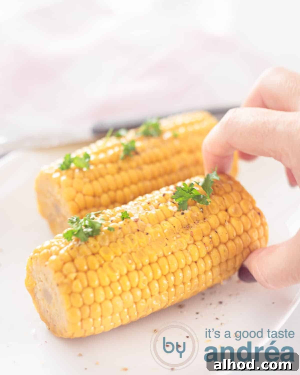 Fire-Kissed Bourbon Butter Corn 5 An appetizing close-up of two bourbon butter grilled corn cobs on a white plate, garnished with parsley. A hand reaches for the front cob, with a soft pink and white towel in the background.