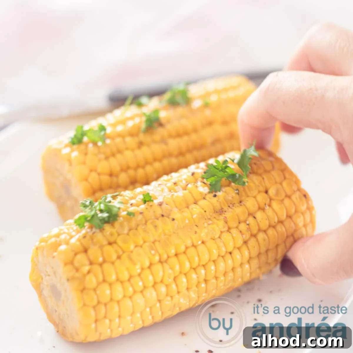Fire-Kissed Bourbon Butter Corn 6 A square photo featuring two buttery grilled corn cobs with fresh parsley on a white background, with a hand reaching for one of them.