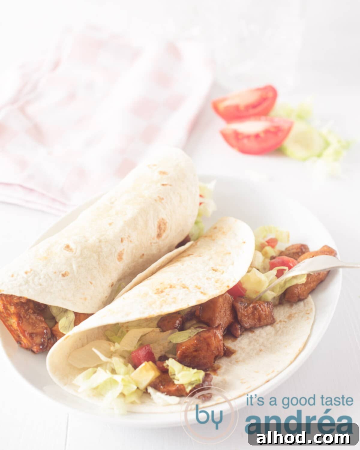 Flavorful Soy Chicken Wraps 2 An elevation photo with a white board. Two wraps on it. Filled with chicken, tomato, lettuce. In the background tomato wedges, lettuce and a pink white checkered tea towel